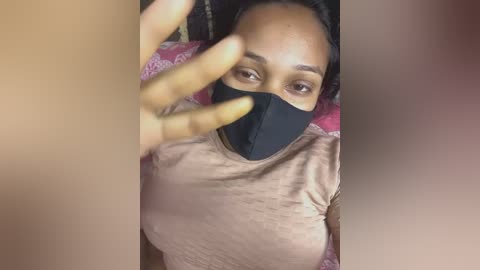 A video of a Black woman with medium skin tone, wearing a black face mask, light brown t-shirt, and pink floral-patterned bedspread.