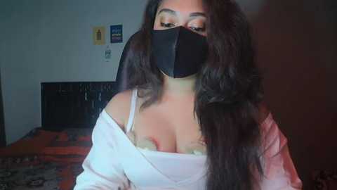 Video of a South Asian woman with long black hair, wearing a black face mask, off-shoulder white top, revealing a green lace bra. Background includes a bed with a patterned blanket, posters on the wall.