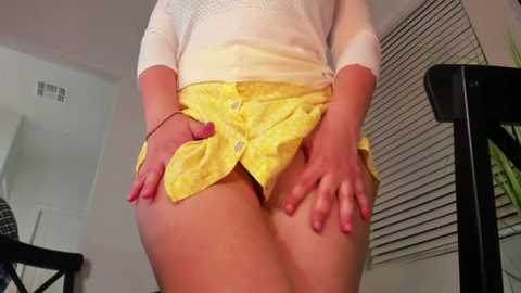 Video of a light-skinned woman in a white top and yellow patterned shorts, exposing her bare buttocks and genital area, standing in a modern, minimally decorated room with a window blind and a chair.