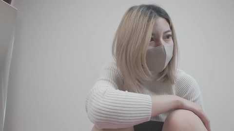 Video of a young Asian woman with straight, shoulder-length blonde hair, wearing a white ribbed sweater, and a white face mask. She's seated with her arms crossed, looking down. Background is a plain, light-colored wall.