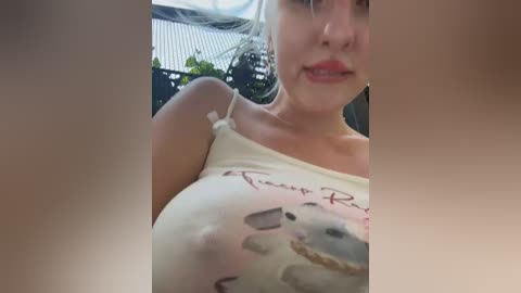 A video of a fair-skinned woman with a slender build, wearing a white tank top with visible cleavage and a playful, slightly mischievous expression, taken indoors with a blurred background.