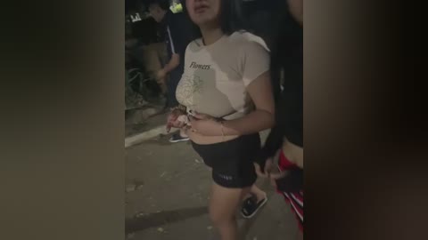 Video of a young woman with a light complexion, medium-length black hair, wearing a white t-shirt and black shorts, walking in a dimly lit urban environment at night.