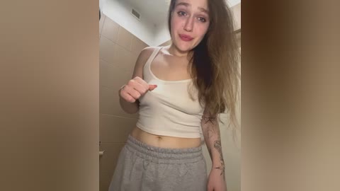 Video of a young woman with light skin and long brown hair, wearing a tight white crop top and loose gray sweatpants, standing in a tiled bathroom, with a playful expression and a raised hand.