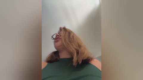 A video of a fair-skinned woman with shoulder-length blonde hair and glasses, wearing a dark green top, looking upwards with a contemplative expression against a plain white background.