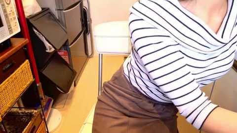 Video of a woman with fair skin wearing a white and black striped blouse and brown skirt, sitting on a white chair in a cluttered, beige-tiled room with a black wicker basket and a black metal table.