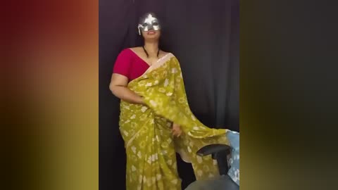 Video of a woman with medium skin tone wearing a red blouse, yellow sari with white floral pattern, silver masquerade mask, black hair, standing in front of a dark background.