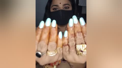 Video of a woman with long, manicured nails, light blue polish, and multiple gold rings. She wears a black face mask and a red bindi. Background is blurred.