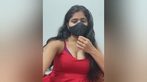 A video of a South Asian woman with long black hair, wearing a red dress, black mask, and purple bra, covering her mouth, standing against a blurred background.
