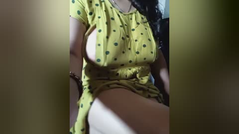 Video of a woman with medium skin tone, wearing a yellow polka-dot dress, showing her left breast. She is sitting with her legs apart, revealing her bare thigh.