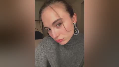 Video of a young woman with light skin, brown hair, and a serious expression, wearing a gray turtleneck and ornate earrings, seated indoors with blurred background.