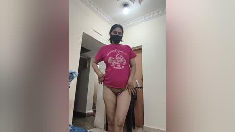 A video of a plus-size woman in a pink t-shirt and black panties standing in a cluttered, beige-walled room with a door, a fridge, and a wooden table in the background.