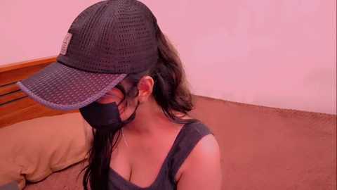 Video of a woman with long dark hair, wearing a black face mask, a black mesh cap, and a low-cut tank top, sitting on a bed with beige sheets and a brown blanket.