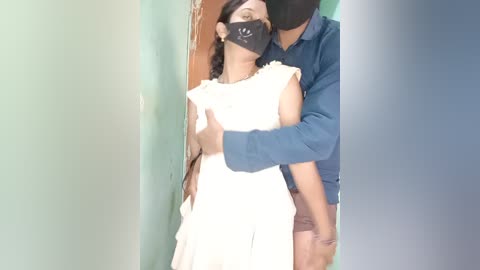 Video of a masked woman in a white dress, being hugged by a man in a blue shirt, standing in a green-walled room.