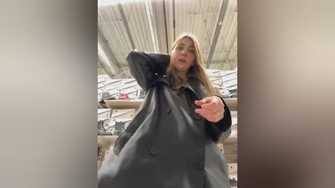 Video of a blonde woman in a dark coat, arms raised, inside a warehouse with metal shelves and industrial ceiling.