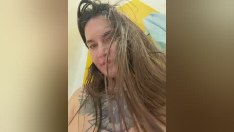 Video of a wet, dark-haired woman with a large, colorful tattoo on her chest, partially visible, in a bathroom with yellow and white towels.