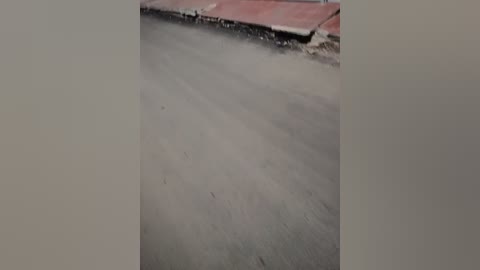 Video of a partially paved street with dirt and scattered debris, showing a rough, unfinished construction scene. The background features a red curb and a concrete sidewalk. The overall scene is dusty and lacks a finished appearance.