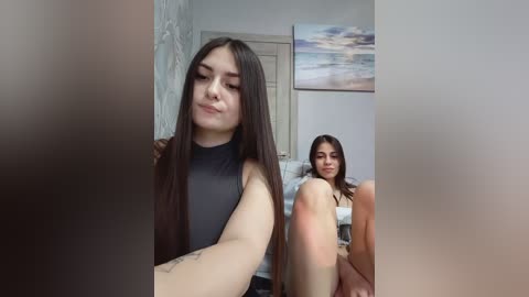 A video of two young women with light skin, long dark hair, and medium build, wearing black tops, sitting indoors near a bed with a beach painting on the wall.