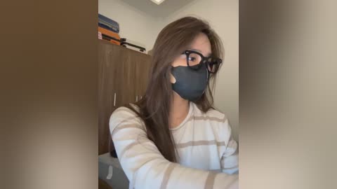 Video of a woman with long brown hair, wearing black-rimmed glasses, a black face mask, and a striped sweater, sitting in a room with a wooden cabinet and stacked blankets.