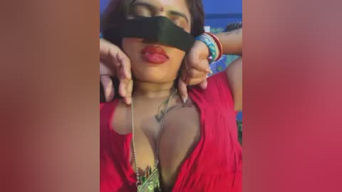 Video of a South Asian woman with medium brown skin, wearing a red top and a black blindfold, holding a green necklace, with a blurred background.