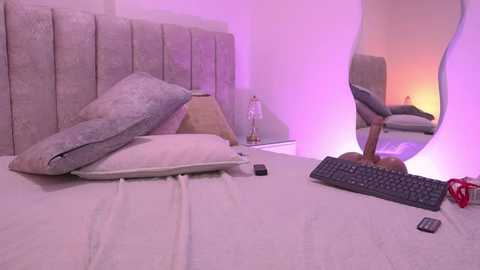 Video of a modern, minimalist bedroom with a light gray upholstered headboard, white bed linens, a decorative pillow, a bedside lamp, a mirror, and a black keyboard on the bed.