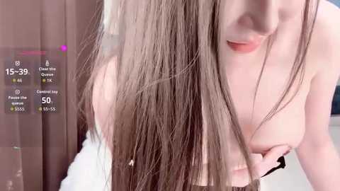 A video of a light-skinned woman with long, straight brown hair, partially covering her small breasts. She's topless, holding her breast with her left hand. The background features a blurred, beige curtain and a digital screen with stats.