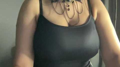 Video of a woman in a tight, black spaghetti-strap tank top, revealing cleavage and a necklace with dangling tassels. Her skin tone is light, and she has a curvaceous figure.
