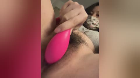 Video of a person with light skin, using a pink vibrator on their pubic hair, while wearing a mask and a floral-patterned shirt.
