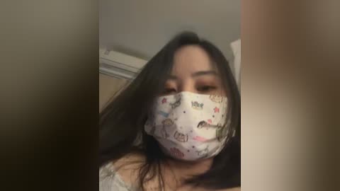 Video of a woman with long, dark hair wearing a white face mask with cartoon designs, standing in a dimly lit room with beige walls and a wooden door in the background.