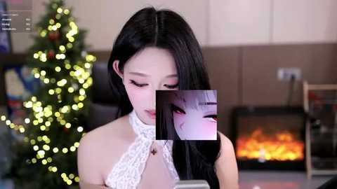 Media: Video of an Asian woman with long black hair and light skin wearing a white lace halter top, standing near a lit Christmas tree and a fireplace, with a close-up overlay of a face with anime-style makeup.