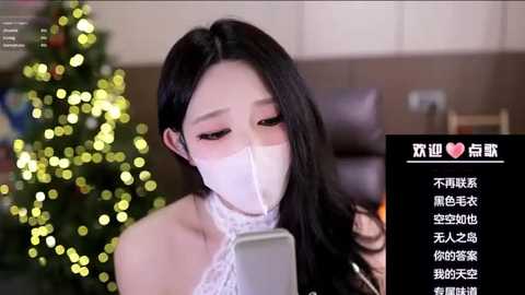 Media: Video of an Asian woman with long black hair wearing a white face mask, seated in a cozy living room with a lit Christmas tree in the background. Text overlay in Chinese.