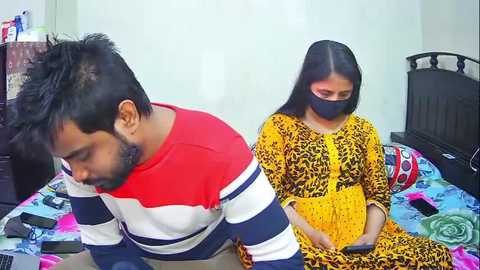 Video of a South Asian couple in a bedroom; man in a red and white striped shirt, woman in a yellow and black leopard print dress, both wearing face masks.