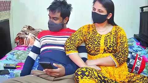 Video of an Indian couple wearing masks, the man in a striped shirt, the woman in a yellow dress, sitting on a colorful bed in a simple room.