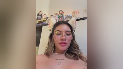 Video of a young, light-skinned woman with long, wavy brown hair, wearing cat ears and large glasses, showing off a heart tattoo on her chest. Background includes a shelf filled with anime figurines.