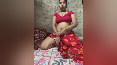 Video of a South Asian woman with medium skin tone, medium build, and long dark hair, sitting cross-legged on a colorful quilt in a rustic room with brick walls. She wears a red top and floral-patterned skirt.