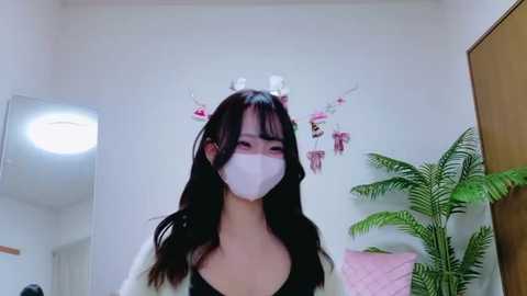 Video of an East Asian woman with long black hair, wearing a white face mask, black top, and a pink headband, standing in a room with white walls, a mirror, a potted plant, and hanging decorations.