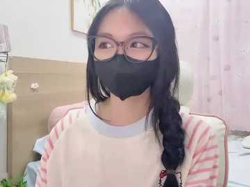 Video of an East Asian woman with long black hair, wearing a pink striped sweater, black face mask, and glasses, sitting indoors with a wooden wall and floral wallpaper in the background.