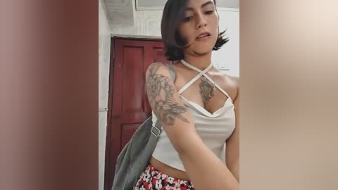 Video of a young woman with short black hair, medium skin tone, wearing a white crop top and red floral skirt, with a black handbag slung over her shoulder.