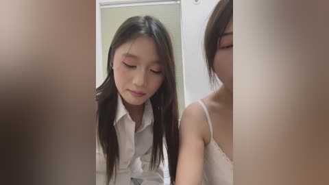 Video of two young East Asian women with long black hair and fair skin, one in a white shirt, the other in a white tank top, standing in a softly lit room with beige walls and a window with blinds.