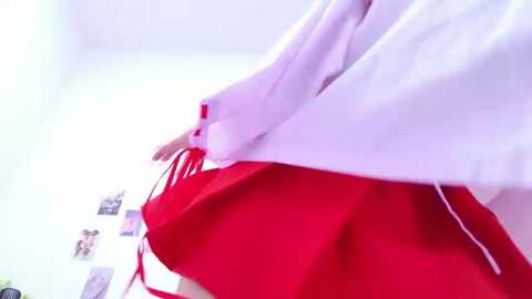 Video of a person in a bright pink kimono with red accents, holding a red obi, against a white wall adorned with photos and a small potted plant.