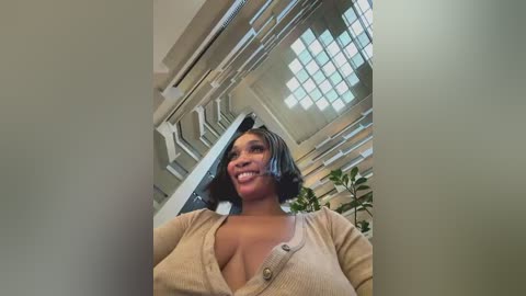 Video of a smiling, slender Black woman with medium skin tone and short, straight black hair, wearing a beige cardigan and a low-cut top, standing in a modern, well-lit, glass-walled building with large windows.