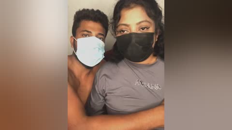 Video of a South Asian couple wearing face masks, smiling and hugging closely, in a dimly lit room. The man has short curly hair, and the woman has curly hair and is wearing a gray shirt.