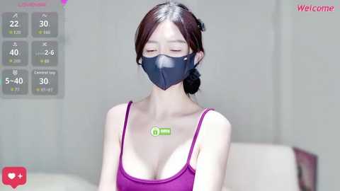 A video of an Asian woman with fair skin and long brown hair, wearing a purple tank top and a dark face mask, standing indoors.