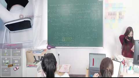 Video of a classroom with a green chalkboard, students, and teacher in a red sweater, wearing a face mask.