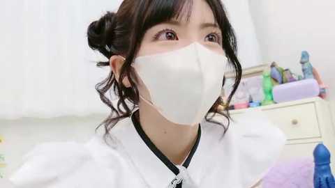 Video of an Asian woman with light skin, wearing a white surgical mask, black hair in a bun, white lab coat, in a brightly lit, white-walled room with a purple storage box and colorful toys in the background.