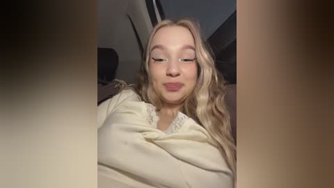 Video of a young woman with long blonde hair, fair skin, and a slight smile, wearing a cream-colored sweater. She is in a car with the window down, partially visible through the frame.
