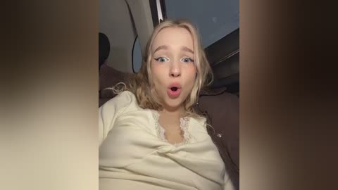 A video of a blonde woman with fair skin, wearing a white lace-trimmed top, inside a car, mouth open in surprise, blue eyes, and a surprised expression.