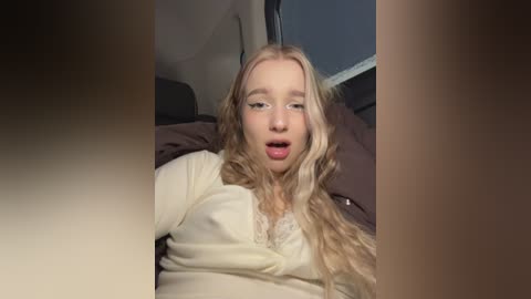 Video of a young blonde woman with wavy hair, wearing a white top, lying on a dark bed, looking surprised and mouth open.