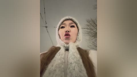 Video of an Asian woman with fair skin and long black hair, wearing a white knit hat and a brown coat, making a kissy face.