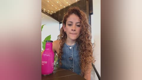 Video of a young girl with curly brown hair, wearing a denim sleeveless top, seated at a counter, holding a pink \"Lusty\" bottle. Background includes a wooden ceiling, plants, and a blurred white wall.