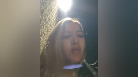 Video of a young woman with long blonde hair, wearing a dark jacket, standing against a textured wall in daylight, with a bright light source behind her.